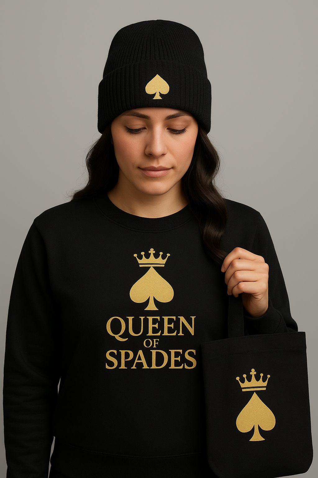 Eine Frau trägt eine schwarze Beanie-Mütze mit goldenem Pik-Symbol, dazu ein schwarzes Sweatshirt mit goldener Aufschrift „QUEEN OF SPADES“ und ein schwarzes Stoff-Tote-Bag mit goldfarbenem Pik-Emblem samt Krone. Neutraler Studio-Hintergrund, klare und kontrastreiche Darstellung.