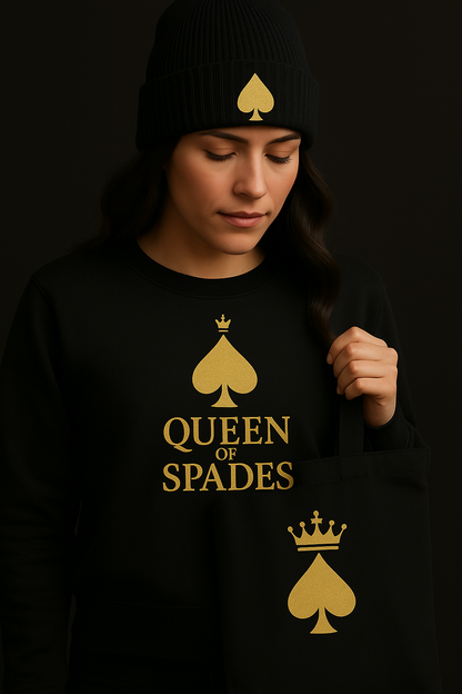 Eine Frau trägt eine schwarze Beanie-Mütze mit goldenem Pik-Symbol, dazu ein schwarzes Sweatshirt mit goldener Aufschrift „QUEEN OF SPADES“ 