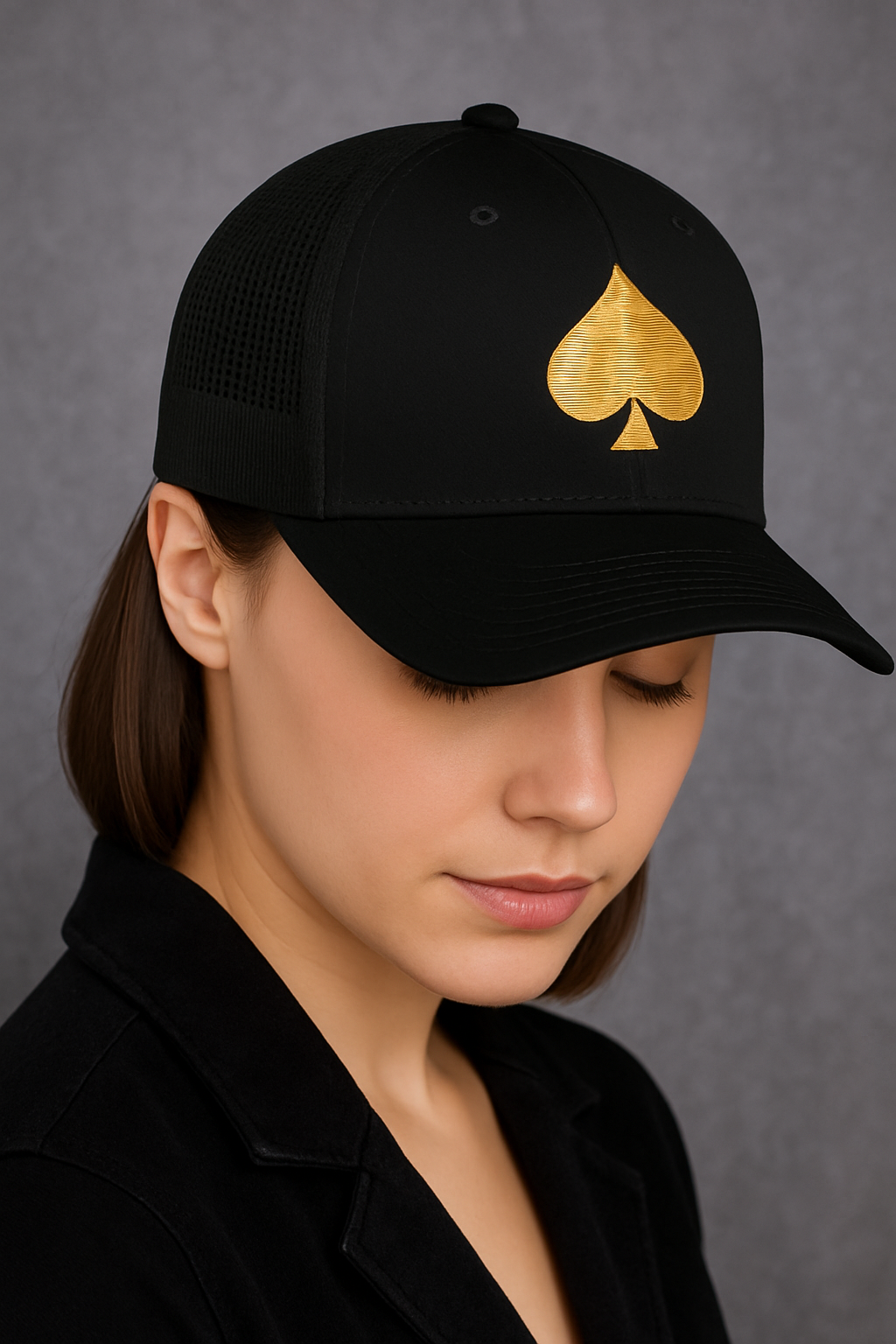 Frau mit schwarzer Trucker-Cap, goldenes Pik-Symbol auf der Vorderseite, Kopf leicht nach unten geneigt.